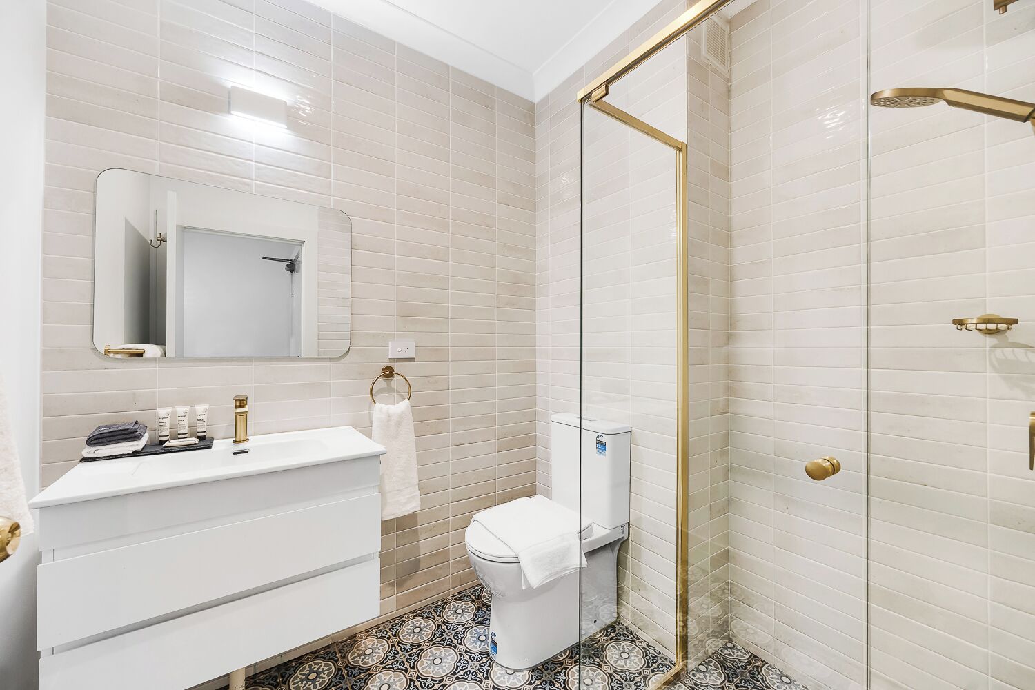 A bathroom with a white sink, mirror, and toilet, a glass shower enclosure, and patterned floor tiles.