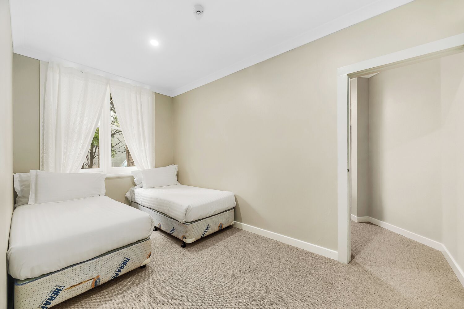 A bedroom with two beds, a window with sheer curtains, a door, and a carpeted floor.