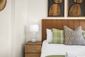 A neatly made bed with patterned pillows, a wooden nightstand with a white lamp, and two wooden wall art pieces above the bed. - Kiahuna 2 (uMhlanga)
