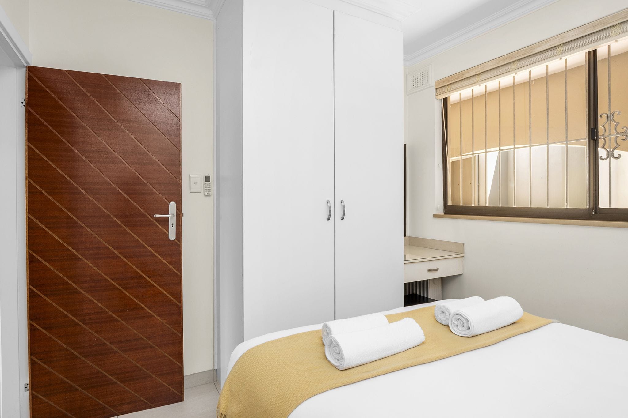 A hotel room with a wooden door, white wardrobe, a bed with a yellow blanket, and a window with blinds.