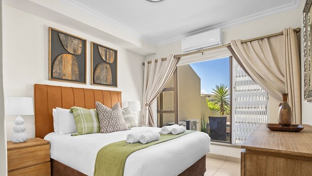 A hotel room with a bed, a nightstand, a desk, a window with curtains, and artwork on the wall. - Kiahuna 2 (uMhlanga)