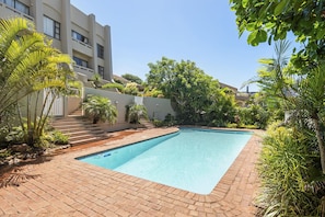 A swimming pool with a brick border, surrounded by greenery and a building with multiple balconies. - Kiahuna 2 (uMhlanga)