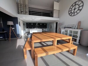 A spacious living area with a wooden dining table, a kitchen in the background, a balcony, and a large wall clock.