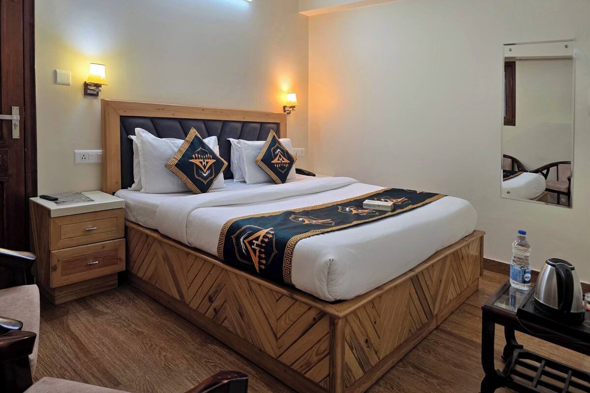 A hotel room with a wooden bed frame, two bedside tables, a nightstand, a mirror, and a small table with a kettle and water bottle.