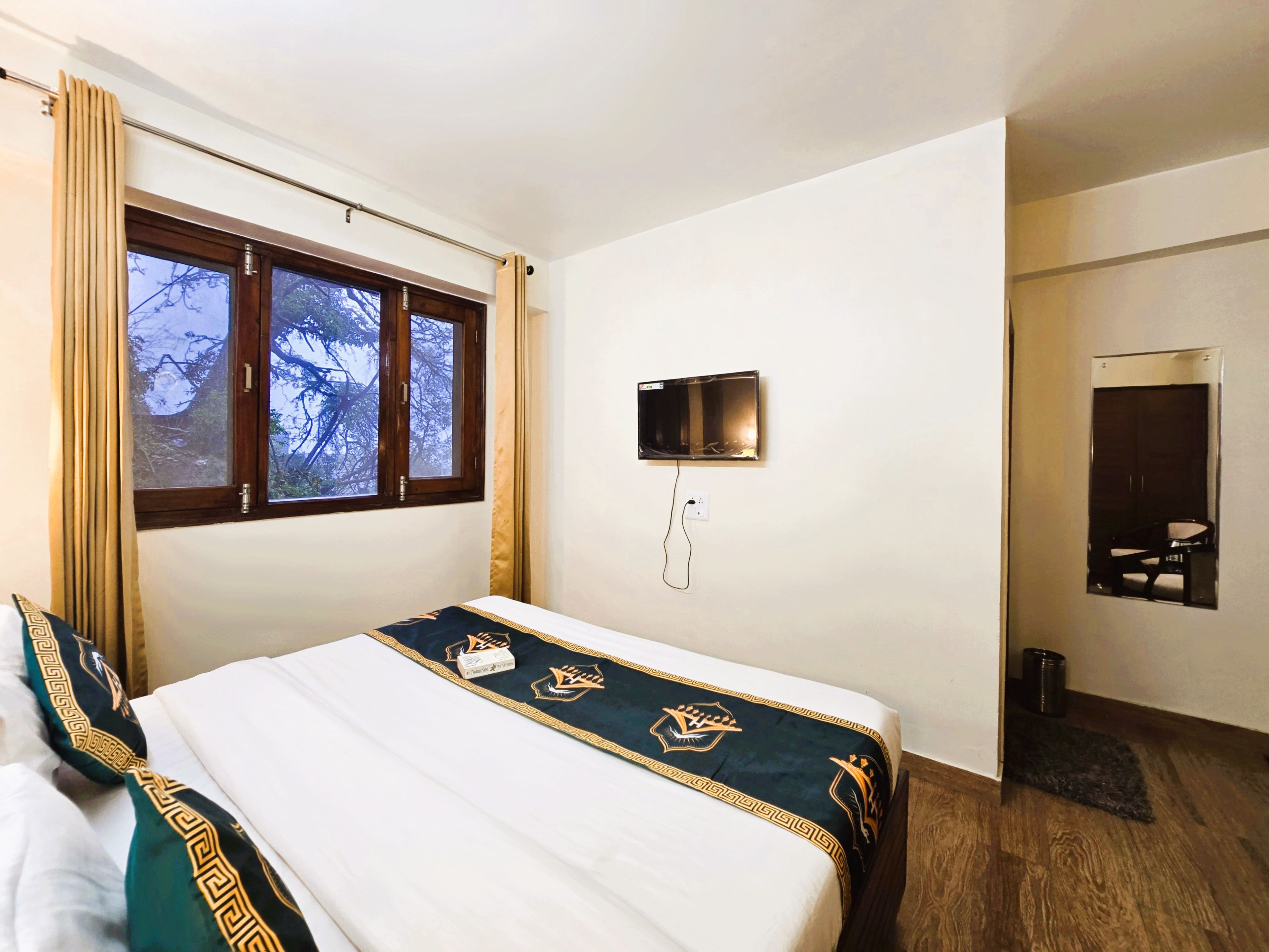 A hotel room with a bed, a window with curtains, a wall-mounted TV, and a door with a mirror.