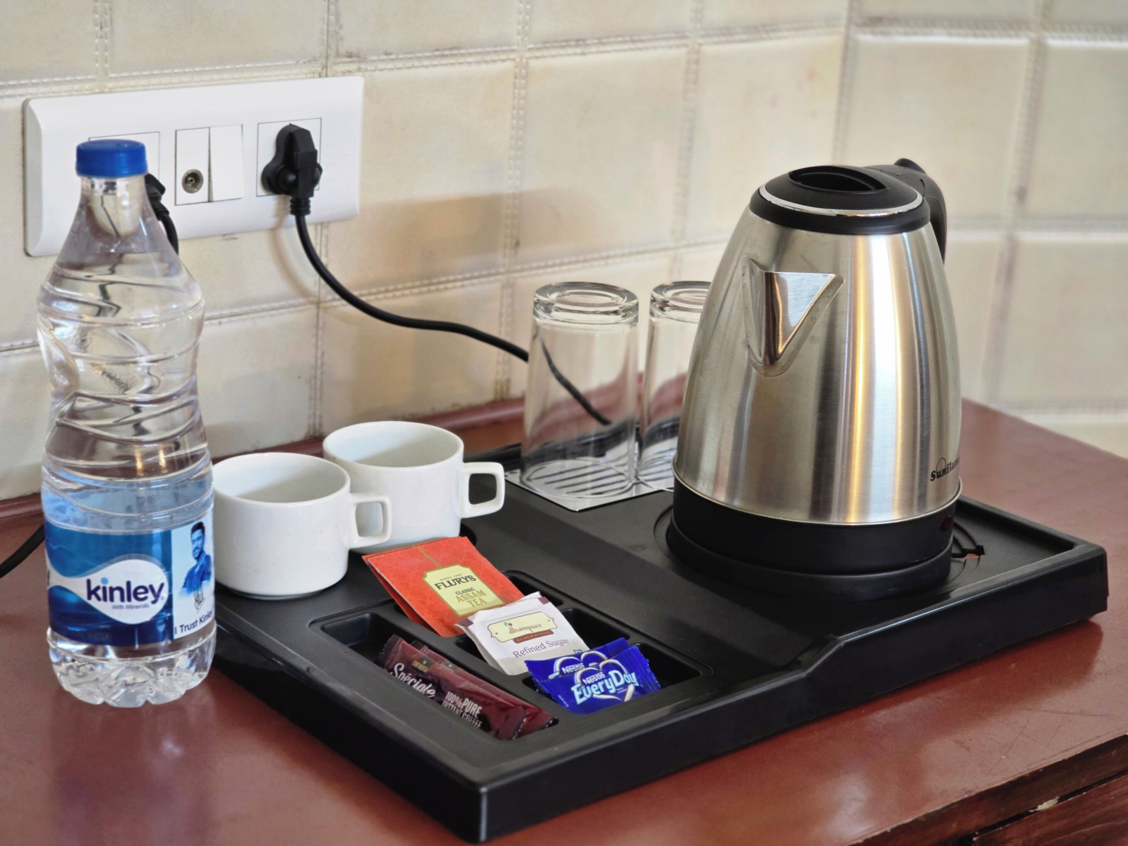 A tray with a water bottle, two white cups, a glass carafe, a kettle, and some snacks.