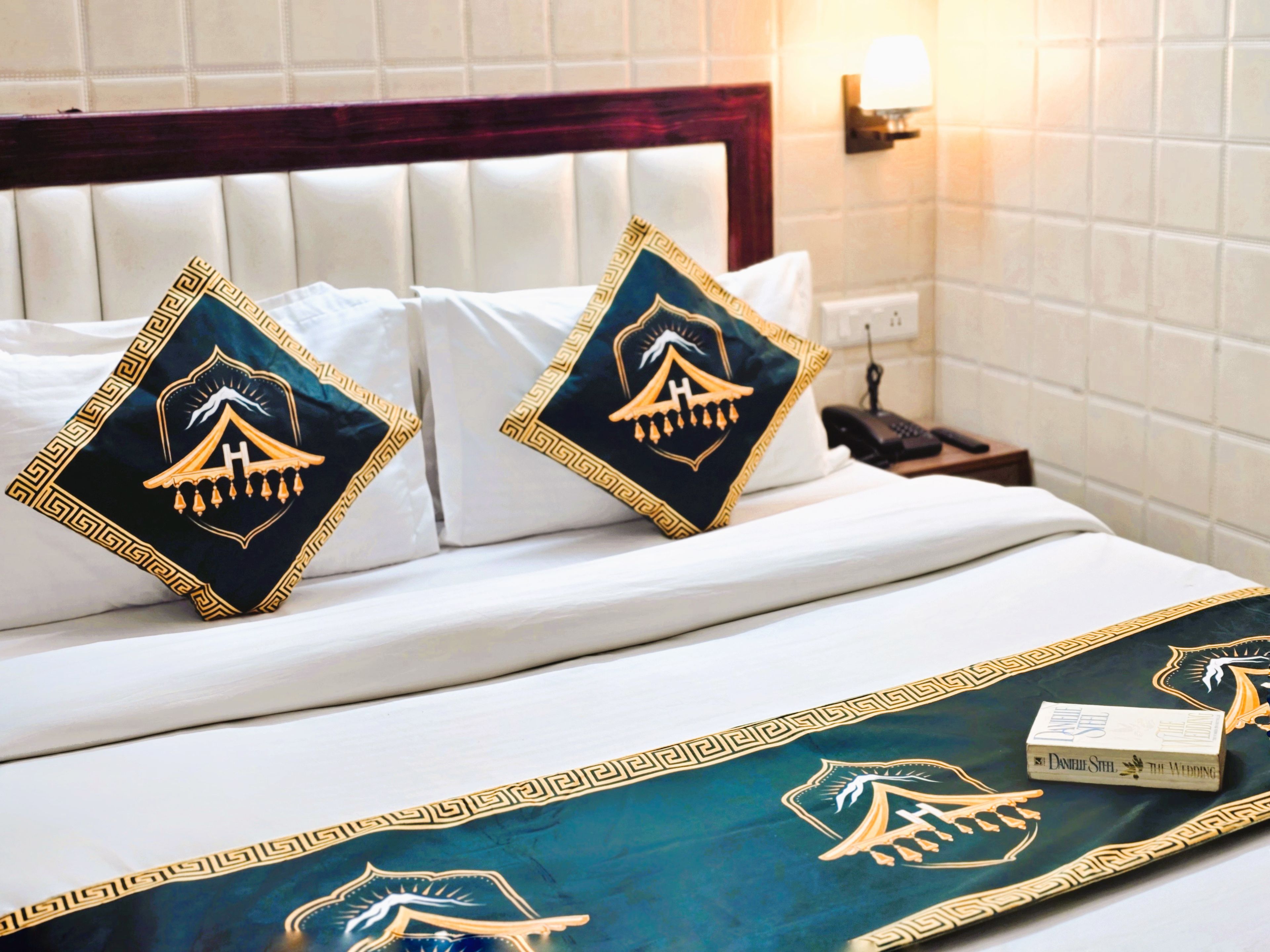 Habitación de hotel con una cama bien hecha, dos almohadas decorativas, una mesita de noche con lámpara y un aplique en la pared.