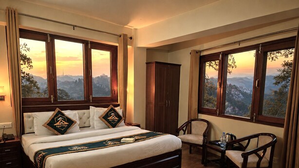 A bedroom with a large bed, two chairs, a wardrobe, and a view of a mountainous landscape through large windows. - Kavin Uphar - The Mall (Shimla)