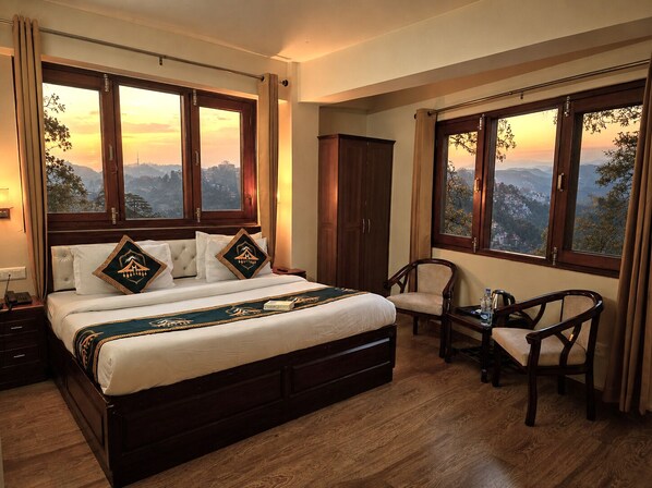 A bedroom with a large bed, two chairs, a wardrobe, and a view of a mountainous landscape through large windows.
