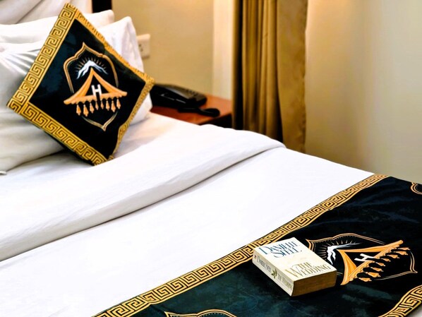 A hotel room with a neatly made bed, a pillow with a crest design, and a book titled 'The Wedding' on the bed. - Kavin Uphar - The Mall (Shimla)
