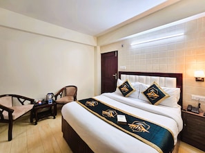 A hotel room with a bed, a desk, a chair, a nightstand, and a wall-mounted lamp.