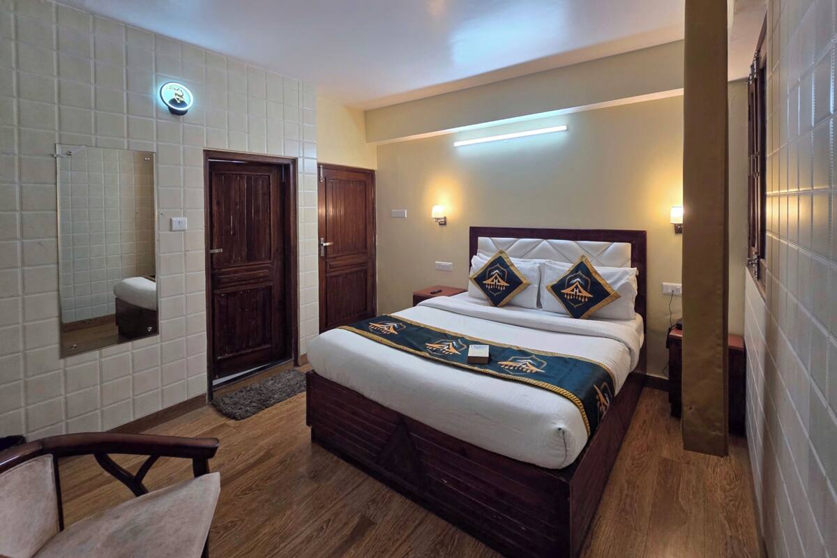 A hotel room with a wooden bed, a bedside table, a chair, a mirror, and a wall-mounted light fixture.