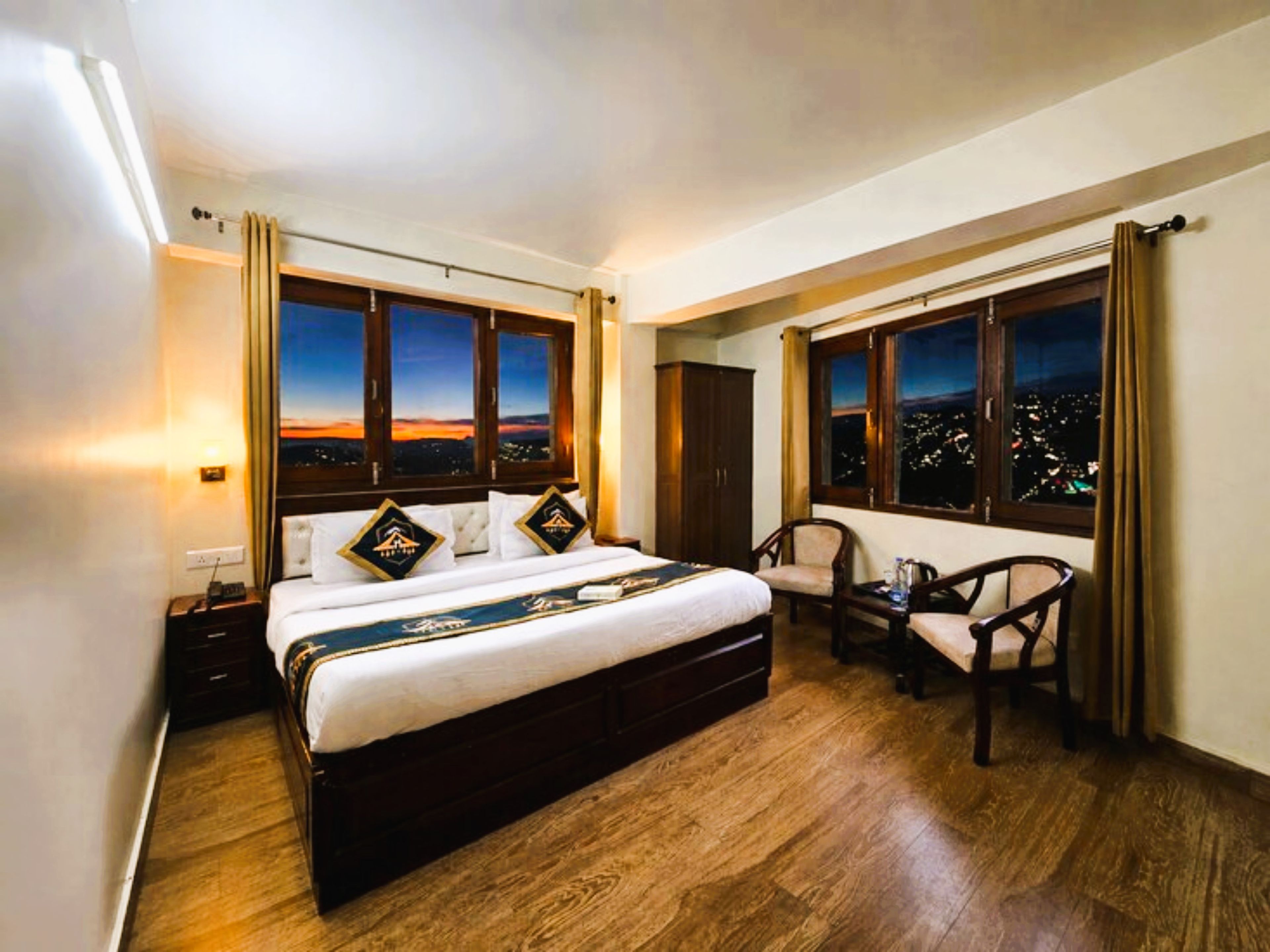 A hotel room with a large bed, a desk, two chairs, and a view of the city at night.