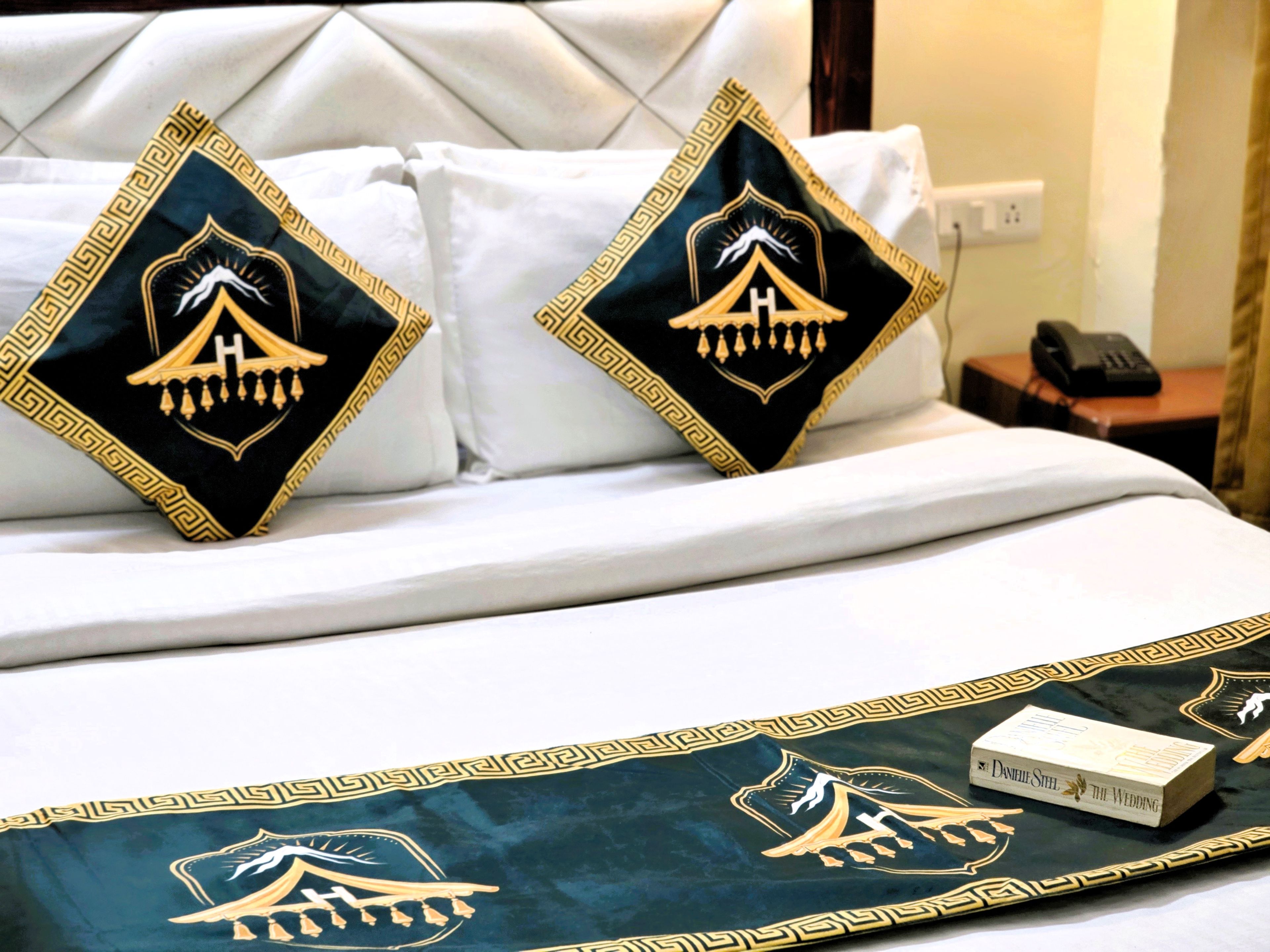 A hotel bed with two decorative pillows featuring a mountain and a tent design, a book titled 'The Wedding,' and a telephone on the nightstand.