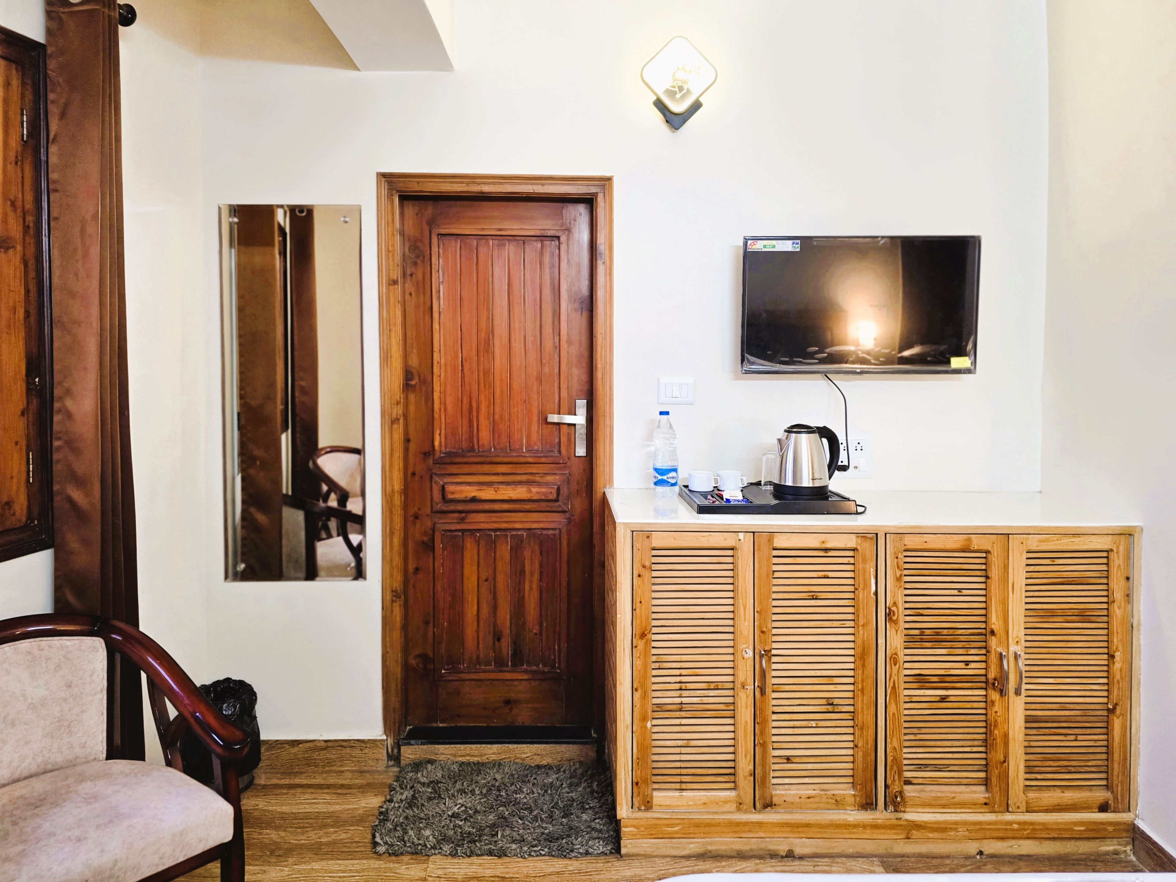 A room with a wooden door, a television mounted on the wall, a cabinet with louvered doors, and a chair.