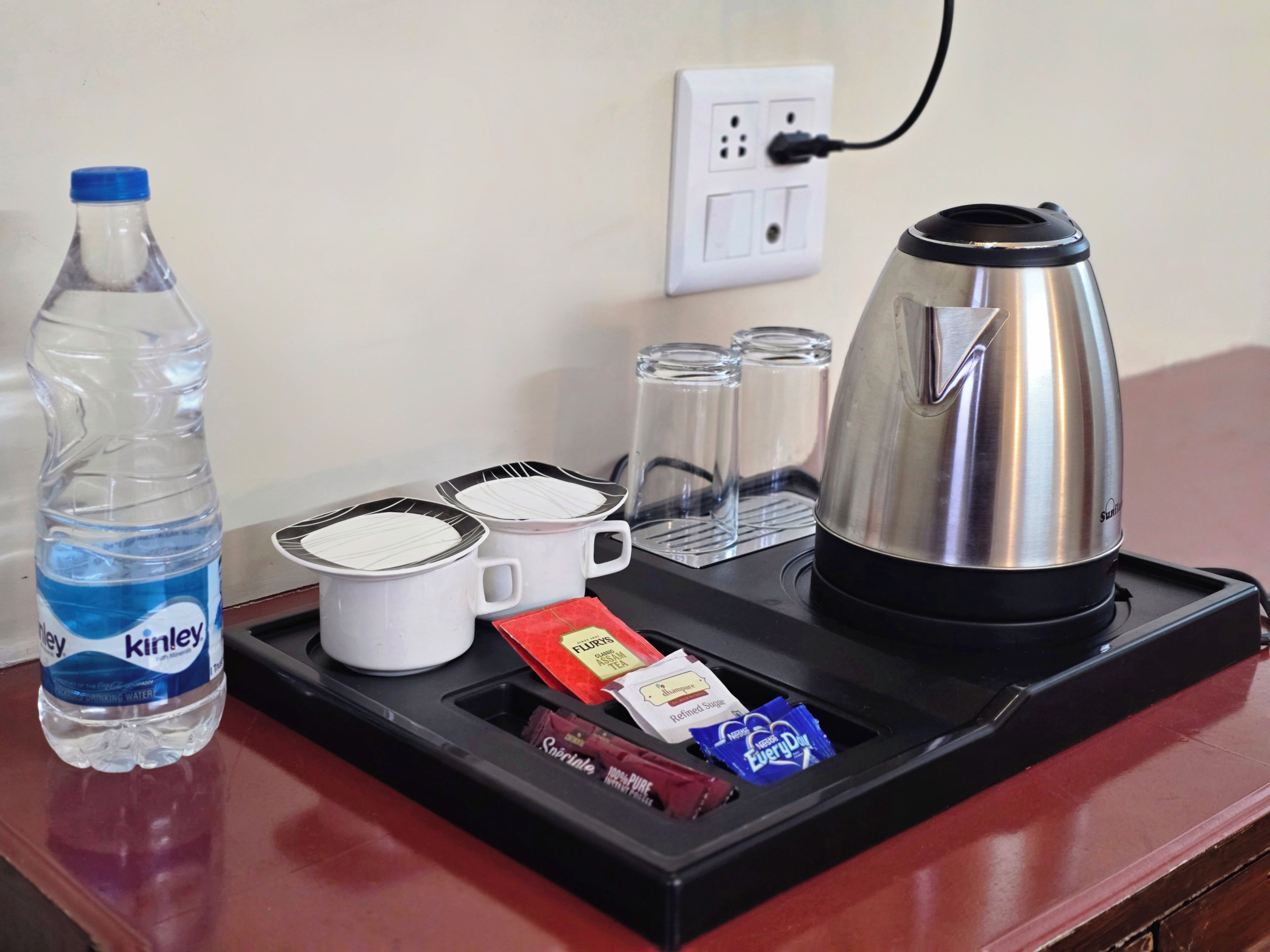 A hotel room's breakfast tray with a water bottle, two mugs, a kettle, glasses, and tea packets.