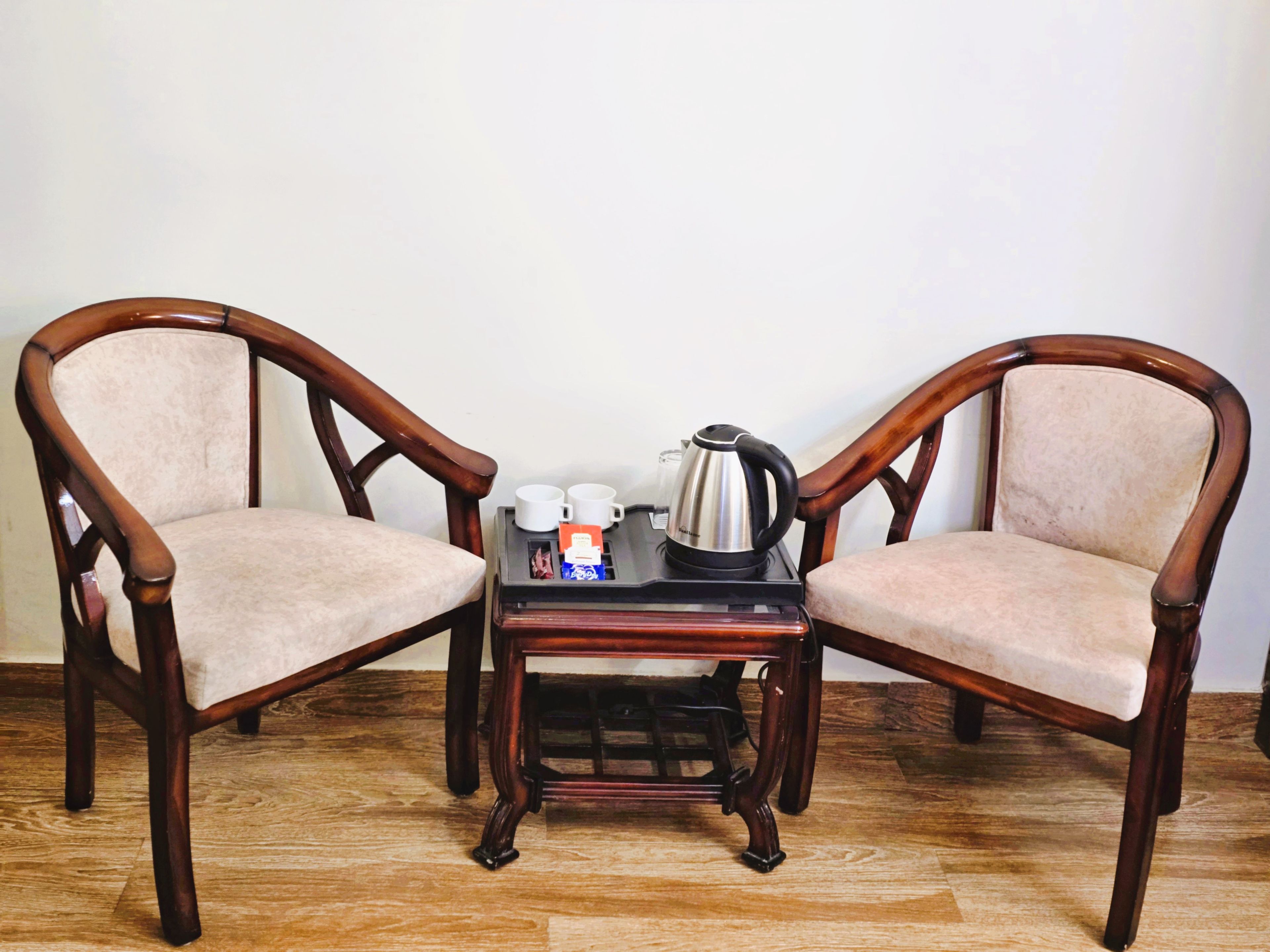 Two wooden chairs with cushioned seats and backs, a small wooden table with a kettle, cups, and a box, against a plain wall and wooden floor.