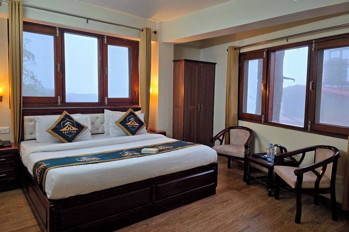 A hotel room with a large bed, wooden headboard, two windows, a wardrobe, a chair, and a bedside table with a lamp.