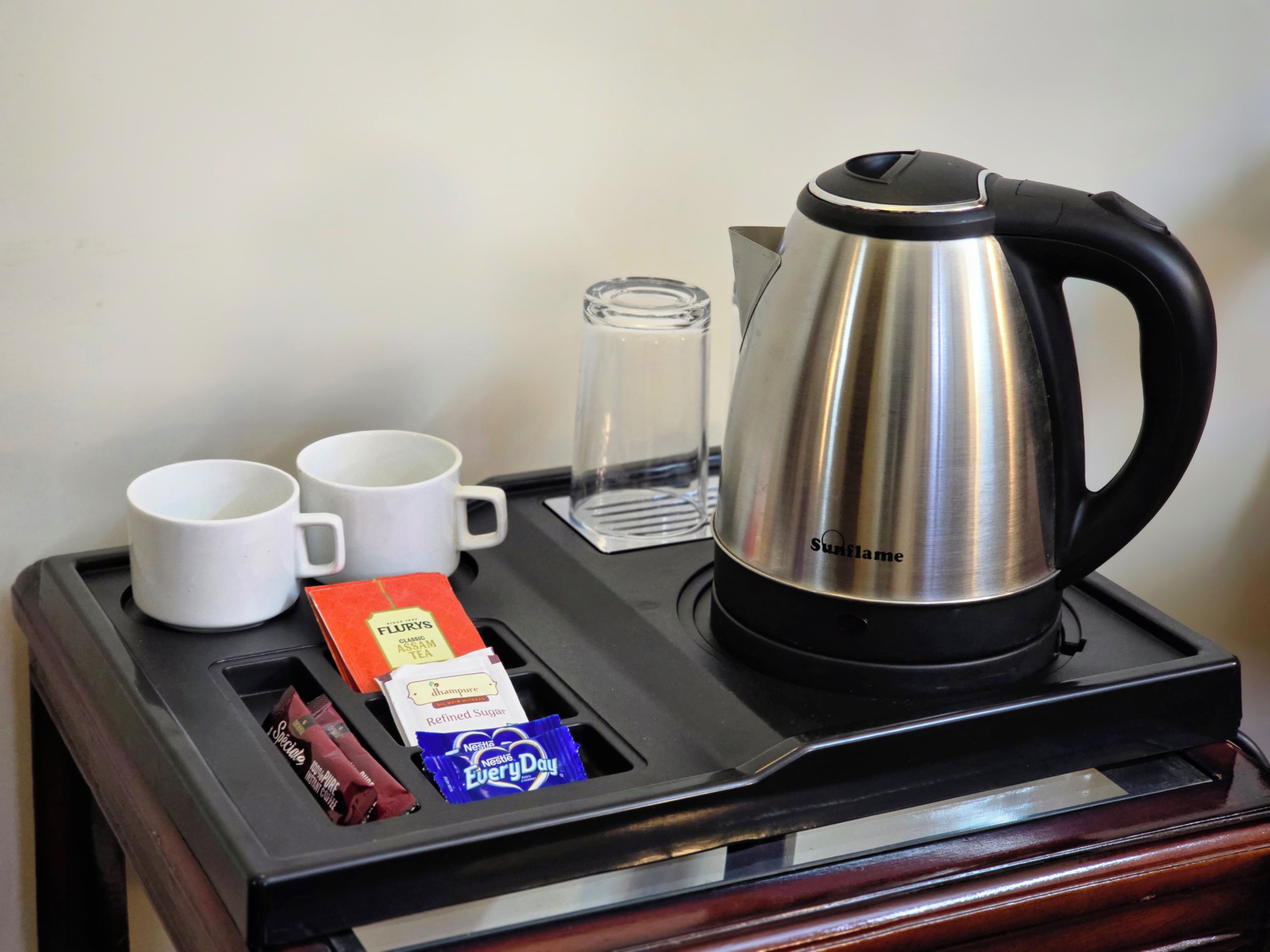 A tray with a stainless steel coffee pot, two white mugs, a glass, and packets of tea and snacks.