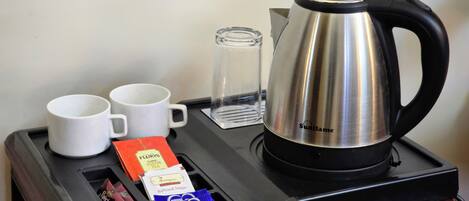 A tray with a stainless steel coffee pot, two white mugs, a glass, and packets of tea and snacks.