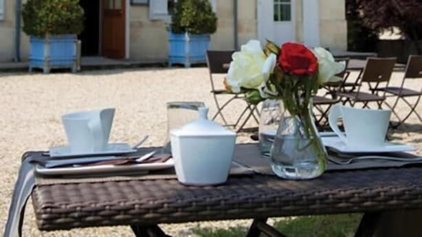 An outdoor dining area with a table set for two, a building with a stone facade, and a door with a sign above it. - Domaine Rollan de by (Bégadan)