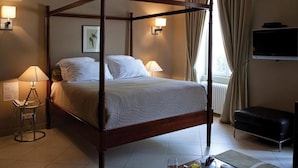 A four-poster bed with a white bedspread, bedside lamps, a flat-screen TV, a black leather ottoman, and a window with curtains. - Domaine Rollan de by (Bégadan)