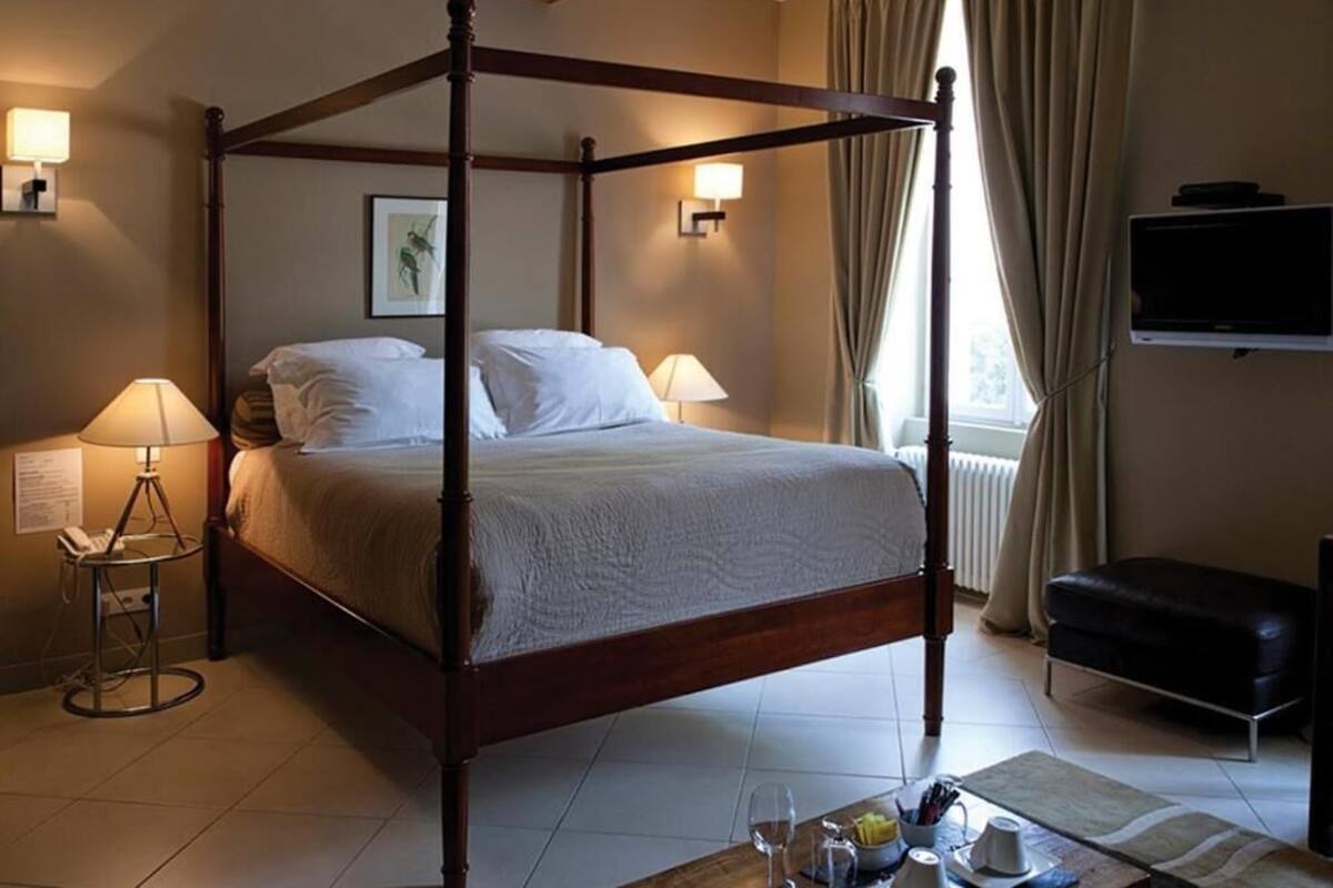 A four-poster bed with a white bedspread, bedside lamps, a flat-screen TV, a black leather ottoman, and a window with curtains.