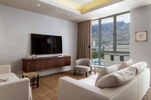 A modern living room with a large flat-screen TV, a sofa, a chair, a small side table, and a view of a cityscape through large windows. - Art Apartment I - Table Mountain View Pool Nomad (Cape Town)