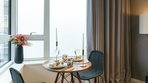 Zona pranzo con tavolo rotondo apparecchiato per due persone, un vaso con fiori e vista sulla città attraverso ampie finestre.