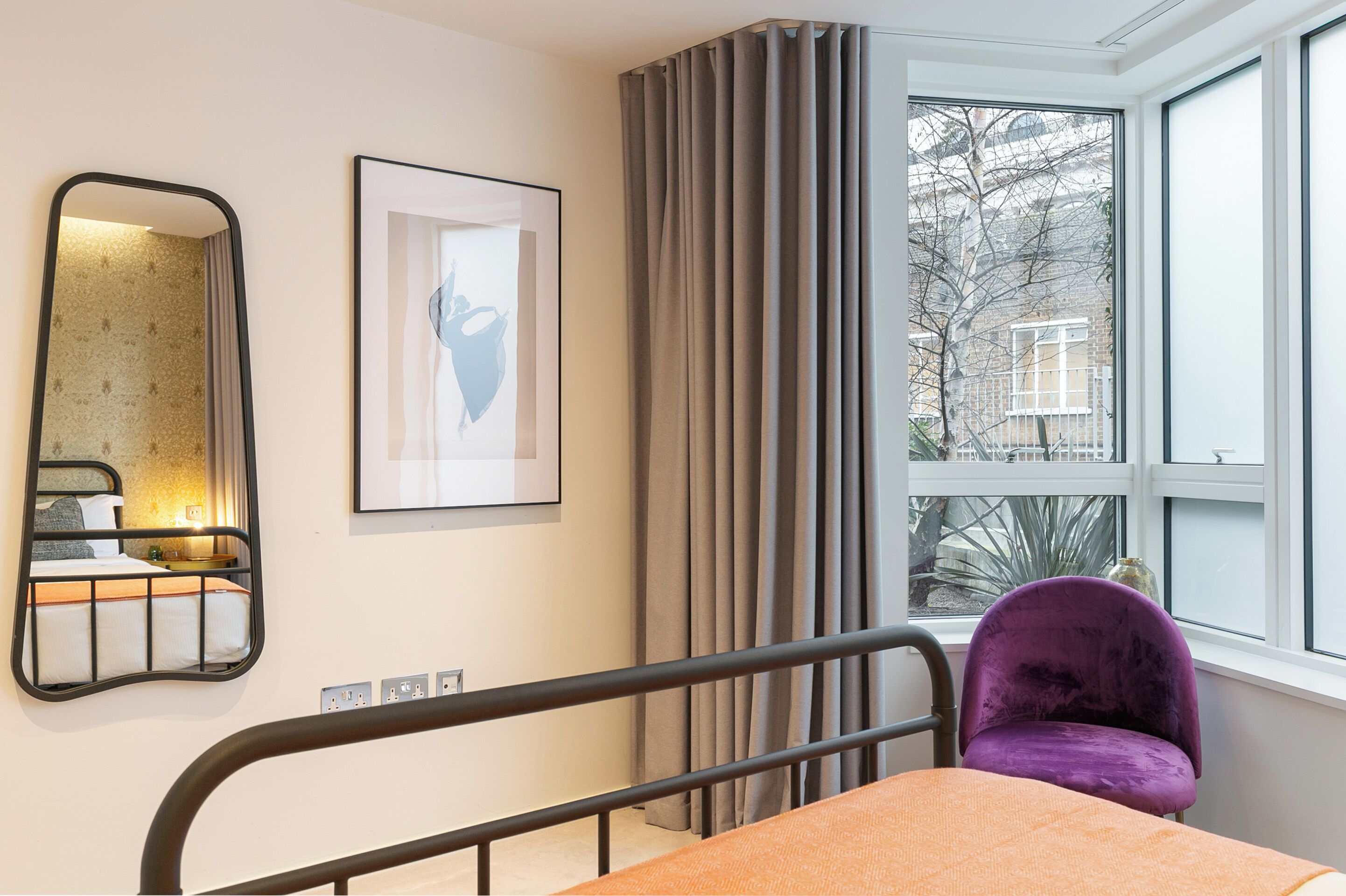 A bedroom with a bed, a chair, a window with curtains, a mirror, and a framed picture on the wall.
