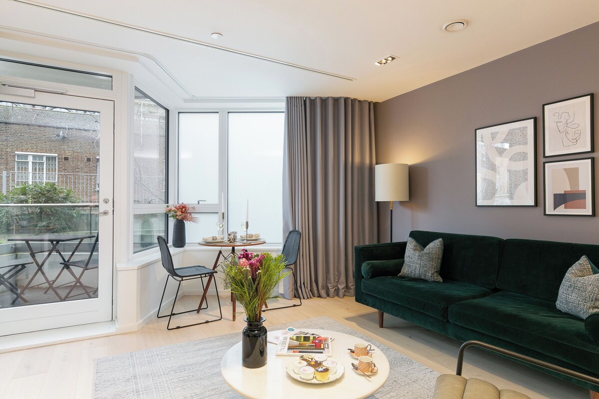 Ein modernes Wohnzimmer mit einem grünen Sofa, einem Esstisch mit Stühlen, einer Vase mit Blumen und einem großen Fenster mit Vorhängen.