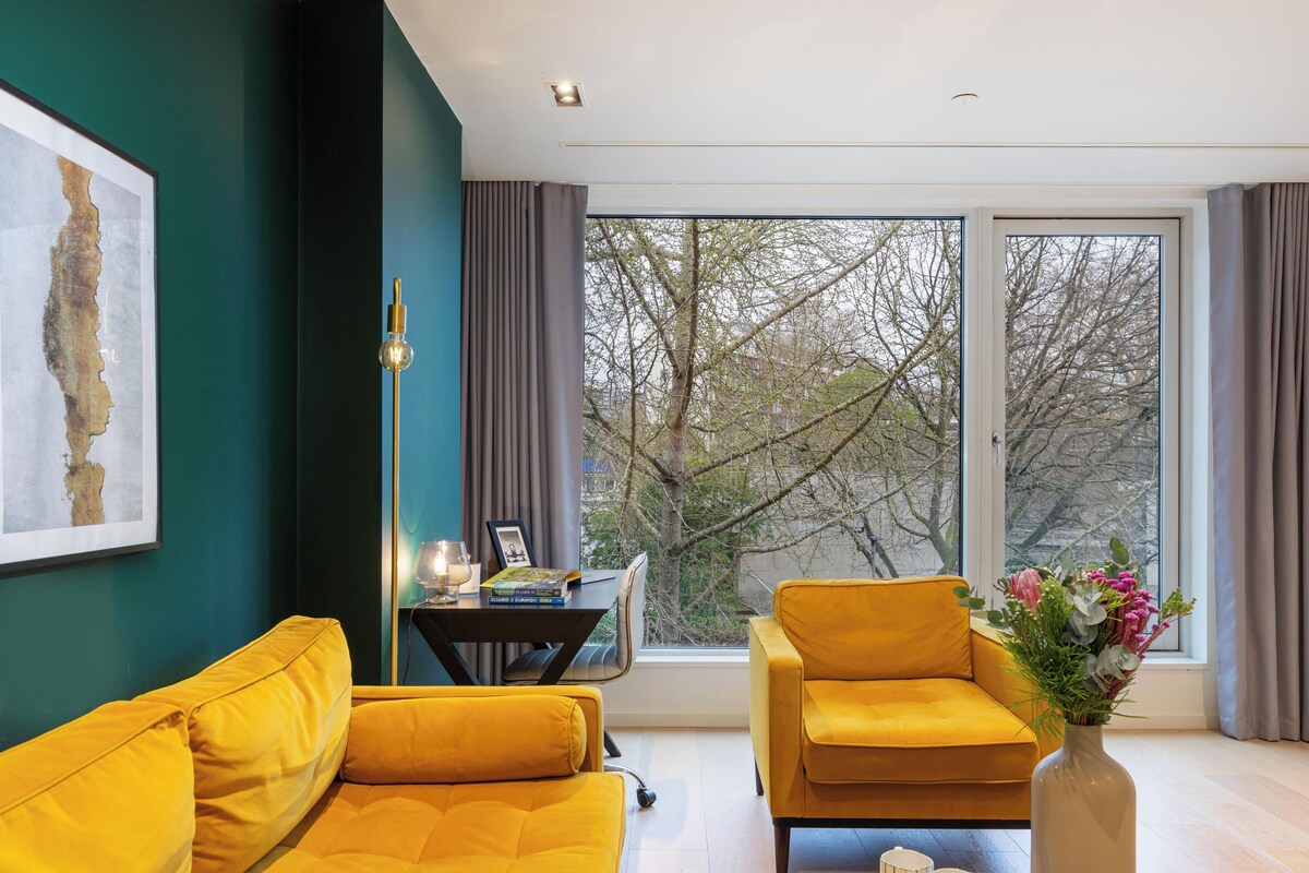 Ein modernes Wohnzimmer mit einem gelben Sofa, einem kleinen Tisch mit einer Lampe und Blick auf Bäume durch große Fenster.