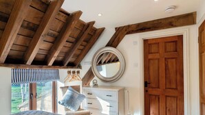A bedroom with a sloped ceiling, wooden beams, a round mirror, a white dresser, a bed, and a wooden door. - Willowberry Barn - Exclusive Home with Hot Tub (Weston Subedge)