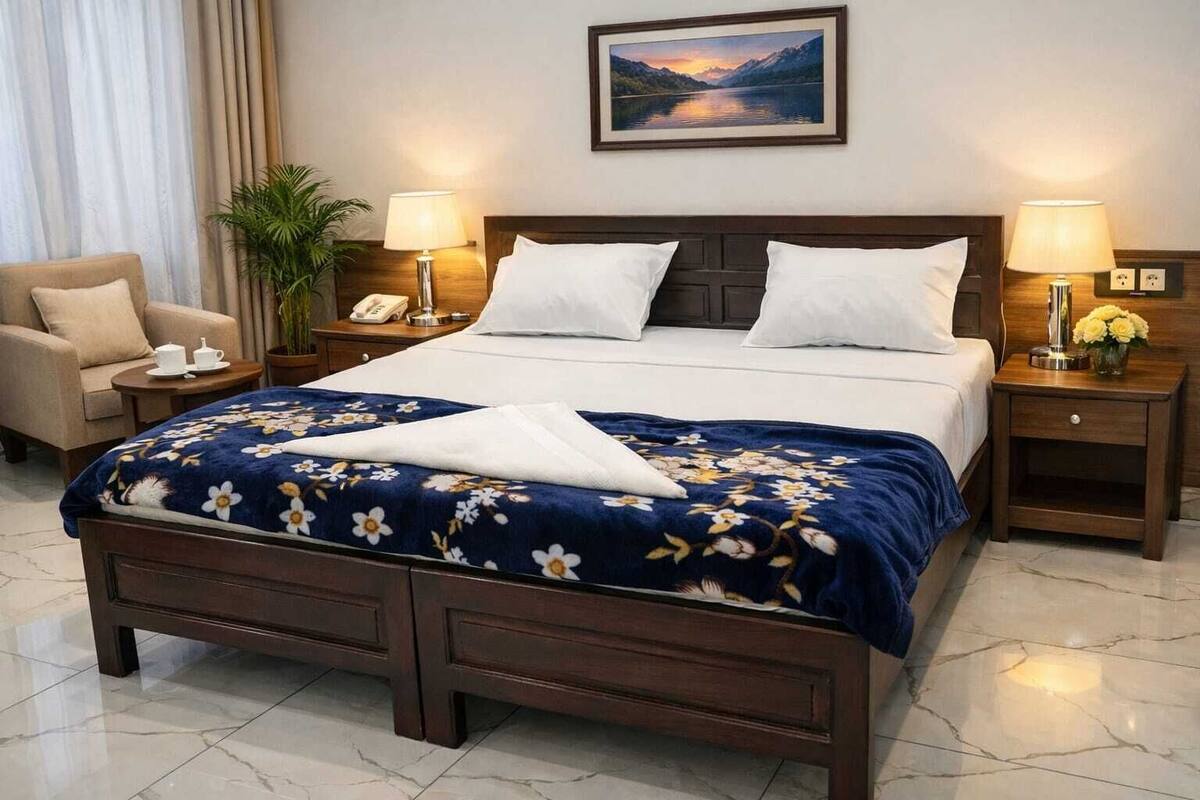 Ein Hotelzimmer mit einem Bett, zwei Nachttischen, einem Sessel, einem Beistelltisch mit Lampe, einem Bild an der Wand und einer Topfpflanze.