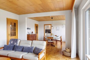 A cozy living room with a sofa, wooden ceiling, dining area with chairs and table, and a kitchen in the background. - Lotterberg Holiday Apartment (Gudensberg)