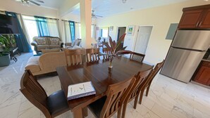 A dining area with a wooden table, chairs, a vase with plants, a sofa, and a kitchen area with a refrigerator. - Serene Family - 4Bed/4Bath Private Ensuite (Cruz Bay)