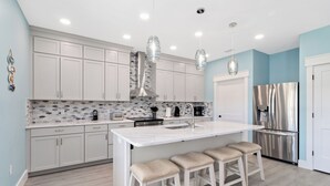 Cocina moderna con armarios blancos, una isla central con taburetes, electrodomésticos de acero inoxidable e iluminación colgante.