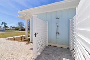 A covered outdoor shower area with a metal hose, a white door, and a gravel pathway leading to a building with blue siding. - Once Upon a Tide – Brand New 2025 Build, Pet Friendly, with Private Pool! (St Joe Beach)