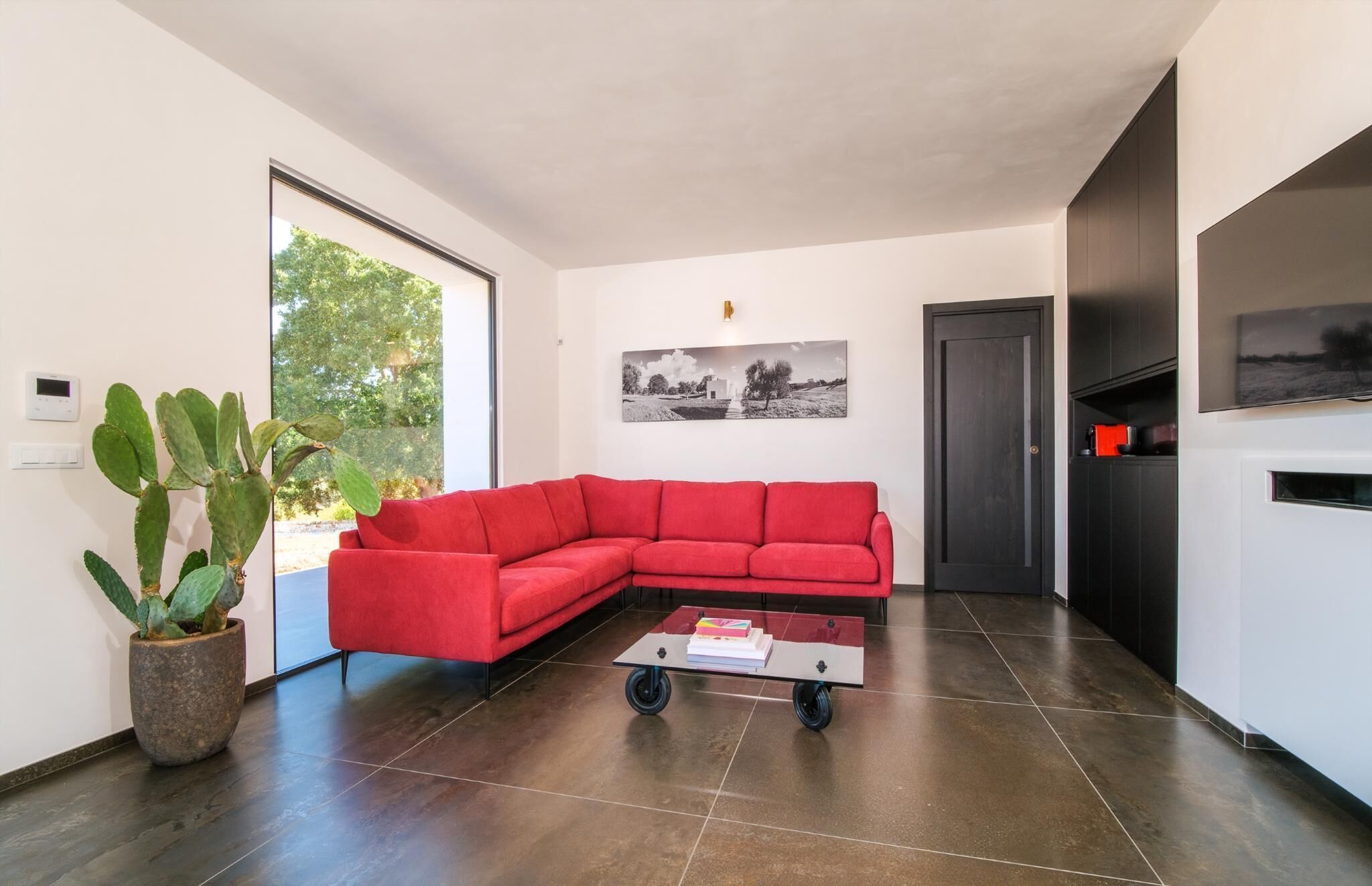 A modern living room with a red sectional sofa, a glass coffee table, a cactus plant, and a door leading to another room.