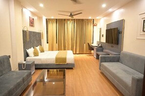 A modern hotel room with a bed, two sofas, a desk, a TV, and a ceiling fan.