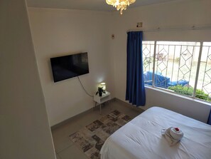 Ein Hotelzimmer mit Bett, Wandfernseher, kleinem Nachttisch mit Lampe, Fenster mit blauen Vorhängen und Blick ins Freie.