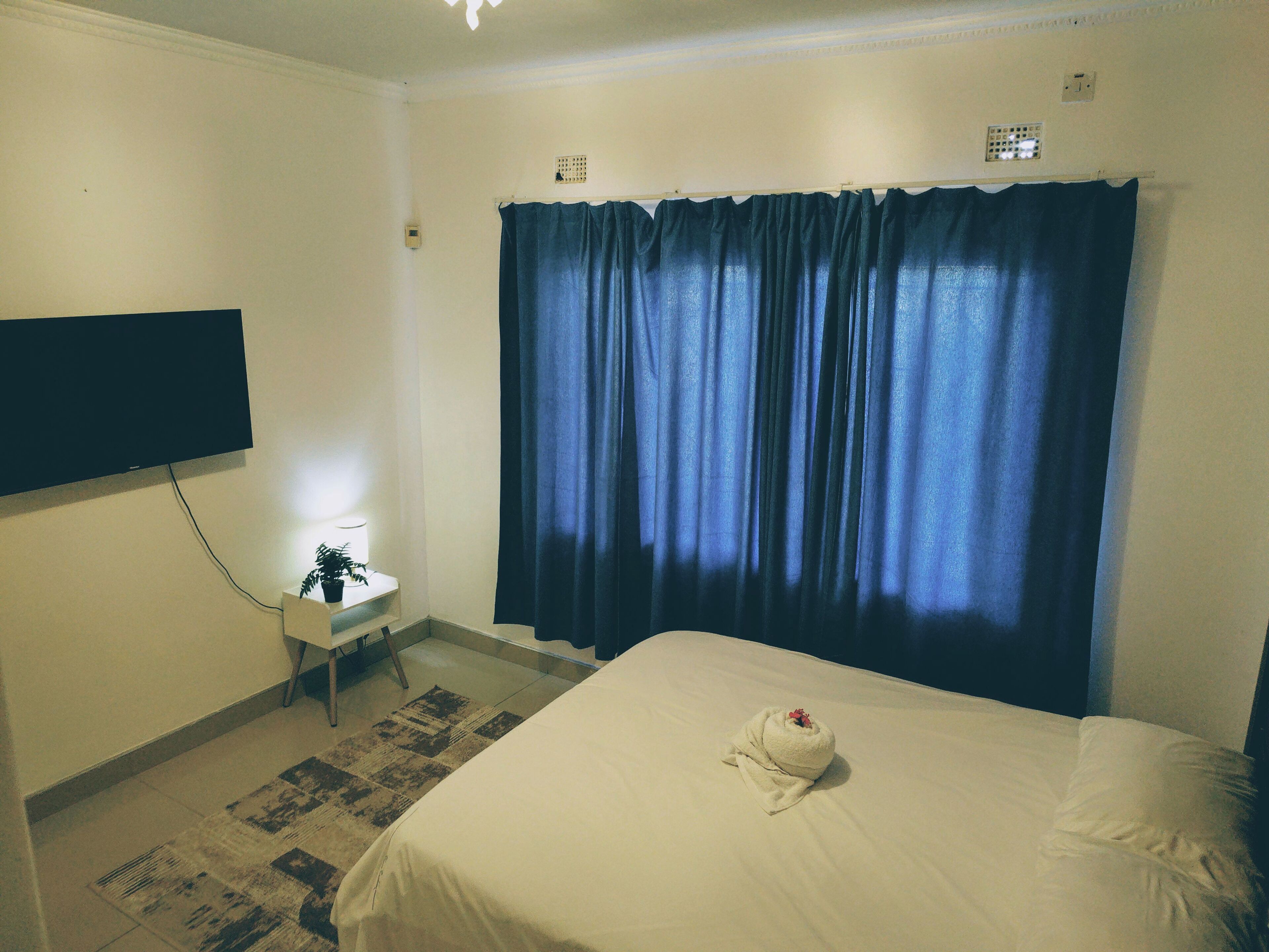 Habitación de hotel con cama, televisor de pantalla plana montado en la pared, mesita de noche con lámpara y ventana con cortinas azules.