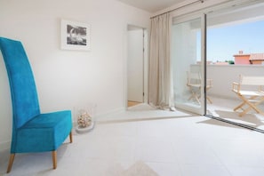 Ein Zimmer mit blauem Sessel, einer Glastür, die auf einen Balkon führt, einem gerahmten Bild an der Wand und einem kleinen dekorativen Gegenstand auf dem Boden.