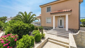 Una casa de dos pisos con una escalera de piedra que lleva a la entrada, rodeada de jardines cuidados y un cielo despejado.