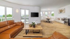 Una sala de estar moderna con un sofá de cuero, una mesa de centro de madera con un jarrón, un televisor de pantalla plana montado en la pared y un comedor con una mesa y sillas blancas.