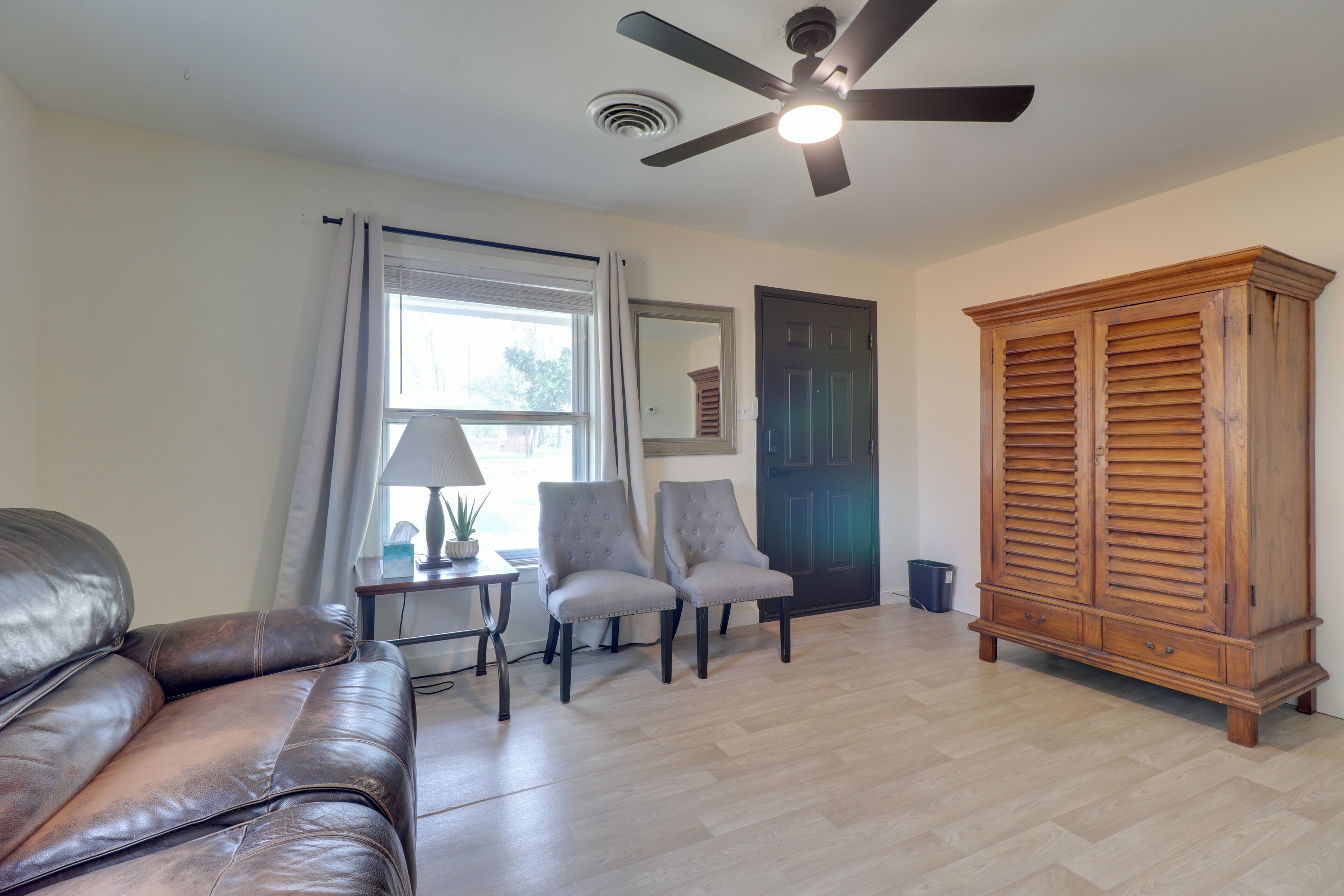 A living room with a leather couch, two armchairs, a wooden cabinet, a ceiling fan, and a window with curtains.