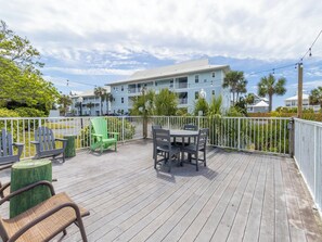Um deck de madeira com móveis de exterior, uma vedação branca e um edifício com varandas ao fundo.
