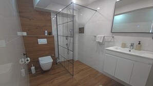 Moderni kylpyhuone, jossa on lasiseinällinen suihku, puulattia, valkoinen pesuallas peilillä ja wc-istuin.
