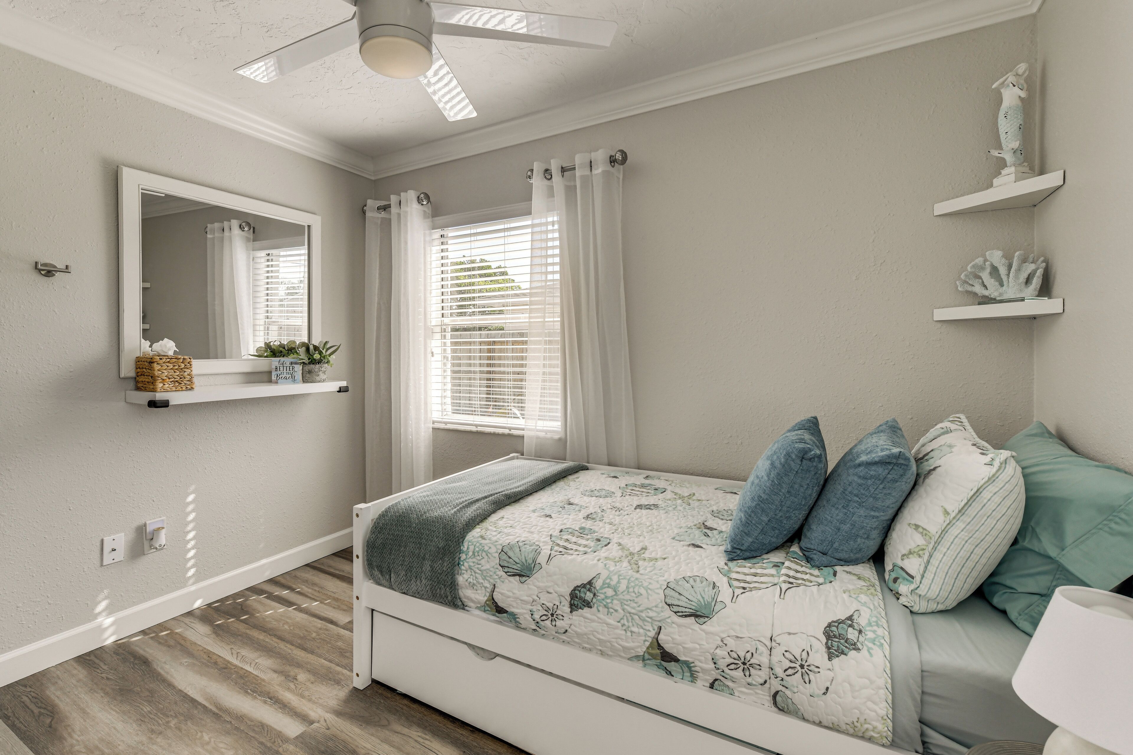 Un dormitorio con una cama, ventilador de techo, ventana con cortinas, espejo y estante con adornos.