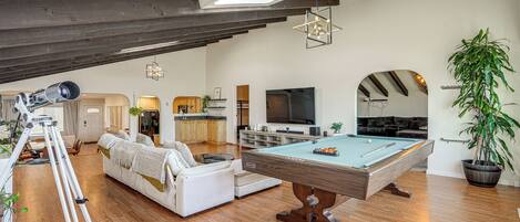 A spacious living area with a pool table, a sofa, a television, and a kitchen in the background.